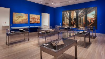 A room in a gallery, walls painted cobalt blue, features items in glass cases and three large, colorful paintings on the walls. Daniel R. Small; "Excavation II," 2016; mixed media installation; installation view at Hammer Museum; courtesy of the artist