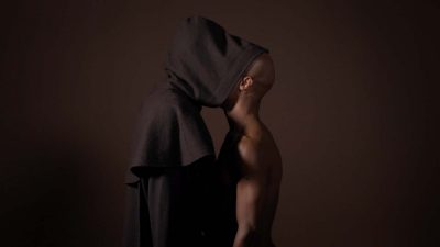 Two male figures stand face to face in front of a dark brown wall. One, a brown man with a shaved head, is shirtless (at right); the other wears a long black hooded robe, the hood portion extending over the face of the man at right. Carlos Motta; "Corpo Fechado-The Devil’s Work," 2018 (detail); installation view; courtesy of the artist and P·P·O·W Gallery