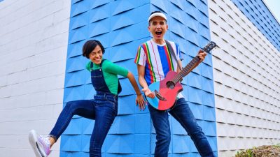 The members of 123 Andrés, a young Hispanic man and woman, wear bright colors and pose animatedly in front of a white and blue building with interesting, textured architecture.
