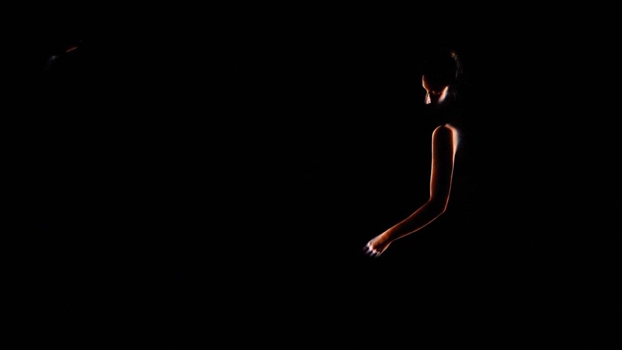 A person's left arm and part of their head is illuminated in darkness, their face still obscured.