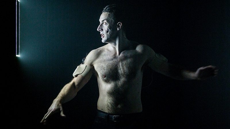 Dancer and theatre artist Andrew Schneider, a white man with dark brown hair, wears a microphone taped to his face, as well as a monitoring device strapped to his arm. His chest is bare, and he looks towards a rectangular light; the rest of the room is dark around him.