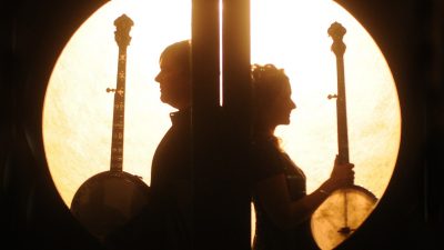 Banjoists Béla Fleck and Abigail Washburn  lean back to back, each holding a banjo vertically, in front of a circular light source.