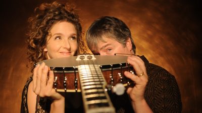 Banjoists Béla Fleck and Abigail Washburn  hold the same banjo, its neck pointing forward, and peer over the edge of the body towards the camera in front of a honey brown background.