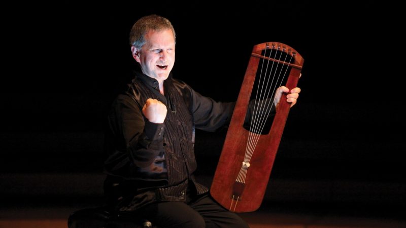 Medieval harpist Benjamin Bagby, a middle aged white man wearing a black shirt and pants, performs "Beowulf" while holding his harp in his lap. He emphasizes the line by making a fist with the other hand.