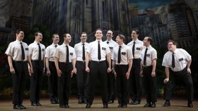 Sam McLellan and company in "The Book of Mormon" North American tour. Eleven white men wear white short sleeved button down shirts, black ties, large dark nametags, and black pants on stage.