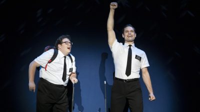 From left, Sam Nackman and Sam McLellan in "The Book of Mormon" North American tour. Both white men wear white short sleeved button down shirt with black pants and ties. One looks more "nerdy" and one raises a fist in the air overhead triumphantly.