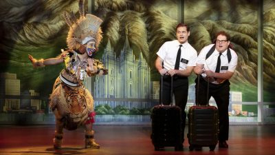 From left, Trinity Posey, Sam McLellan, and Sam Nackman in "The Book of Mormon" North American tour. Posey wears full African tribal garb with colorful face paint and a large headdress. McLellan and Nackman wear white short sleeved button down shirts and black pants and ties as they duck slightly behind their rolling suitcases.