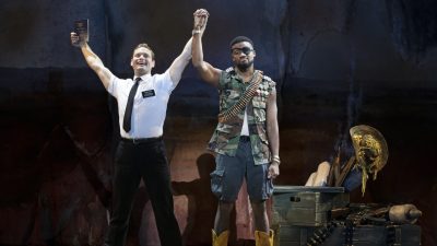 From left, Sam McLellan and Dewight Braxton Jr. in "The Book of Mormon" North American tour. McLellan, wearing a white short sleeved button down and black pants and tie, raises both arms overhead triumphantly. In one hand is a bible; in the other is the hand of Braxton Jr., who is dressed as rebel with a belt of ammo around his neck and an eyepatch. His expression is entirely flat.