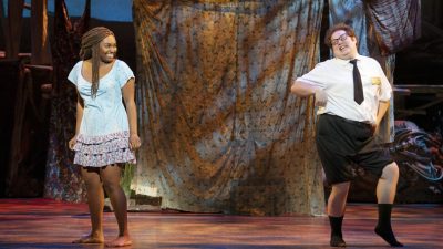 Keke Nesbitt and Sam Nackman in THE BOOK OF MORMON North American tour. Nesbitt, a young Black woman with long braids pulled back into a half up ponytails, wears a light blue dress with three rows of floral ruffles at the hem and looks laughingly towards Nackman, who wears a white short sleeved button down shirt and black tie and pants, which are rolled up to his knees. He strikes a flirtatious pose.