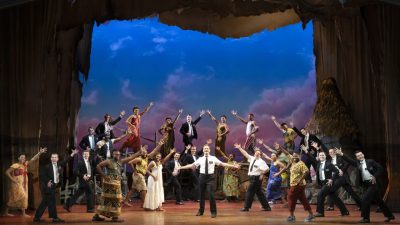 Sam McLellan and company in "The Book of Mormon" North American tour. McLellan, a white man in a white short sleeved button down shirt and black pants a tie, stands in the center of the rest of the cast, who all gesture towards him as if during a grand finale.