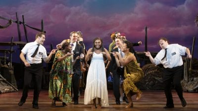 From left, Sam McLellan, Keke Nesbitt, Sam Nackman, and company in "The Book of Mormon" North American tour. Everyone gathers around Nesbitt, a young Black woman with long braids in a white maxi dress; they sing, dance, and point towards her.