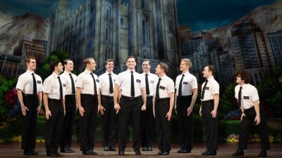 Sam McLellan and company in "The Book of Mormon" North American tour. Eleven white men wear white short sleeved button down shirts, black ties, large dark nametags, and black pants on stage.