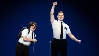 From left, Diego Enrico and Sam McLellan in "The Book of Mormon" North American tour. Both white men wear white short sleeved button down shirt with black pants and ties. One looks more "nerdy" and one raises a fist in the air overhead triumphantly.