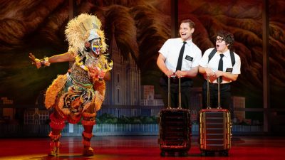 From left, Trinity Posey, Sam McLellan, and Diego Enrico in "The Book of Mormon" North American tour. Posey wears full African tribal garb with colorful face paint and a large headdress. McLellan and Nackman wear white short sleeved button down shirts and black pants and ties as they duck slightly behind their rolling suitcases.