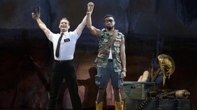 From left, Sam McLellan and Dewight Braxton Jr. in "The Book of Mormon" North American tour. McLellan, wearing a white short sleeved button down and black pants and tie, raises both arms overhead triumphantly. In one hand is a bible; in the other is the hand of Braxton Jr., who is dressed as rebel with a belt of ammo around his neck and an eyepatch. His expression is entirely flat.