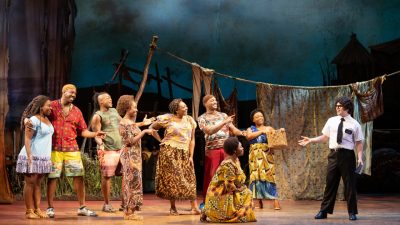 Cast members perform on stage in "The Book of Mormon" North American tour. A group of  11 Black men and women dressed as African villagers look towards one white man in a white short sleeved button down shirt and black pants and tie, holding a bible.