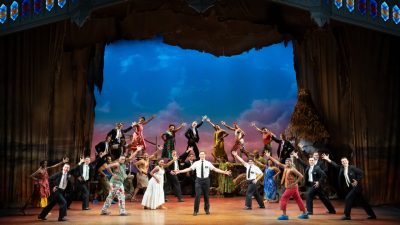 Sam McLellan and company in "The Book of Mormon" North American tour. McLellan, a white man in a white short sleeved button down shirt and black pants a tie, stands in the center of the rest of the cast, who all gesture towards him as if during a grand finale.