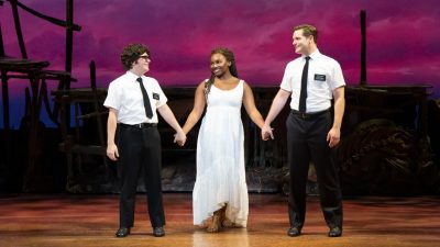 From left, Diego Enrico, Keke Nesbitt, and Sam McLellan in "The Book of Mormon" North American tour. The two white men hold hands with the young black woman, between them, in front of a stage background of a pink sky at sunset.