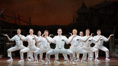 Craig Franke and company in "The Book of Mormon" North American tour. A group of white men in white shirts, pants, and ties, squat low with their arms bent at 90-degree angles.