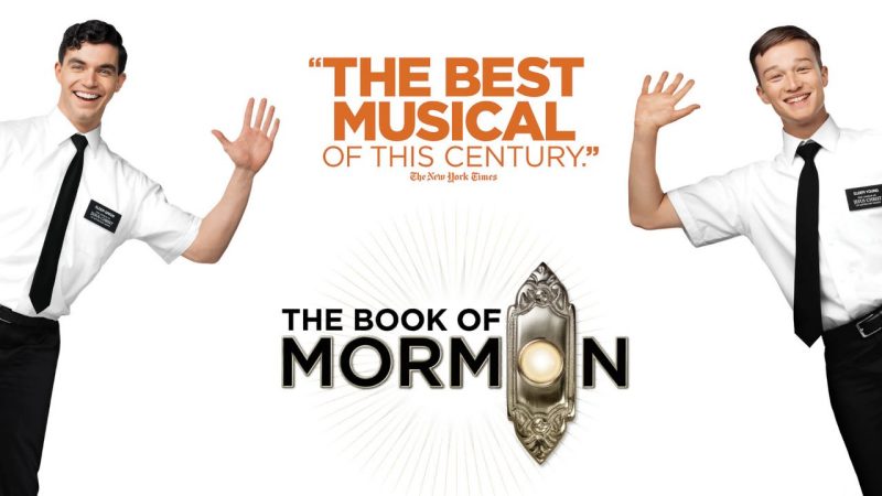 Two white men in white short sleeved button downs and black pants wave and lean into the frame. Between them, orange text on a white background reads "'The best musical of this century.' The New York Times"  The Book of Mormon logo, with the second O in Mormon replaced by a doorbell, is below with a faint starburst pattern surrounding it.