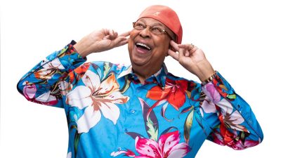 Cuban jazz pianist Chucho Valdes, an older Hispanic man, wears a coral hat and a blue and multicolored floral button down shirt. He smiles widely, his hands on either side of his face.