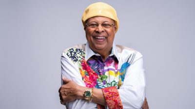 Cuban jazz pianist Chucho Valdes, an older Hispanic man, wears a butter yellow hat and a white and multicolored floral button down shirt. He smiles widely, his arms crossed over his body.