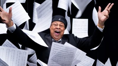 Cuban jazz pianist Chucho Valdes, an older Hispanic man, wears a black Kanga hat and a tuxedo. He smiles widely, hands in the air, as sheets of music rain down all around him.