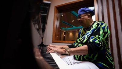 Cuban jazz pianist Chucho Valdes, an older Hispanic man, wears a purple hat and a blue and green patterned jacket. He plays a piano in a recording studio.