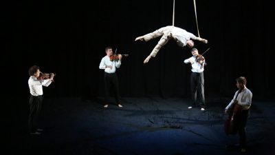 A man wearing all white hangs by a trapeze over the heads of a string quartet, four men in white shirts and black pants.