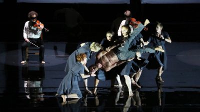 The cast of Circa's "Opus" perform on stage, clustered together and holding a man upside down. In the background, two members of a string quartet play while blindfolded.