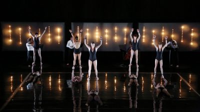 The cast of Circa's "Opus" perform on stage wearing black leotards or pants. Five women stand on the hands of men laying down beneath them, while a string quartet plays in the background.
