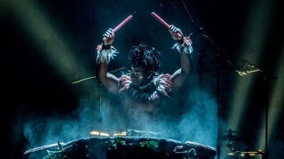 A member of Cirque Kalabanté, a Black man with feathered accessories around his neck and wrists, holds two thick drumsticks aloft while drumming, theatrical fog rolling up from the large drums.
