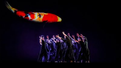 The dancers of Cloud Gate Dance Theatre of Taiwan wear all black and perform on stage, a projection of a Koi fish behind them. Photo by LIU Chen-hsiang