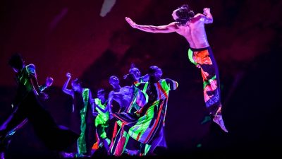 The dancers of Cloud Gate Dance Theatre of Taiwan wear black and neon costumes and perform on stage, a man soaring through the air in the foreground. Photo by LIU Chen-hsiang