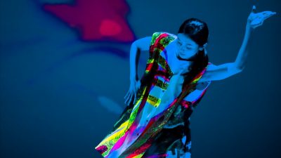 A dancer with Cloud Gate Dance Theatre of Taiwan performs on stage wearing a neon costume that glows under a black light. She holds her arms elegantly. Photo by LEE Chia-yeh