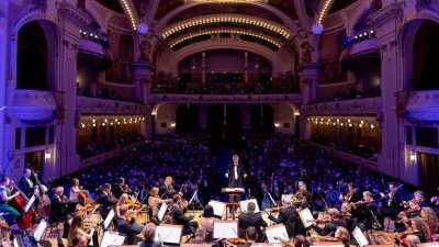 The Czech National Symphony Orchestra performs on stage, dim blue lights cast on the hall and audience behind them.