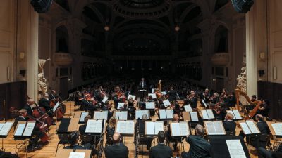 The Czech National Symphony Orchestra performs on stage in a grand concert hall, the auditorium dark behind them.