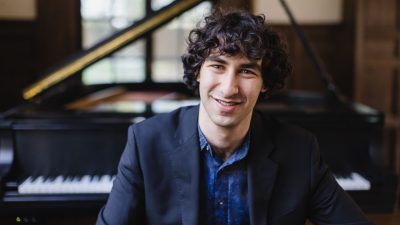 Pianist Maxim Lando, a young white man with medium length curly brown hair, wears a dark blazer over a denim button down shirt. Behind him, a grand piano is out of focus.