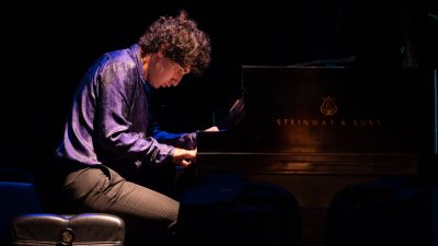 Pianist Maxim Lando, a young white man with medium length curly brown hair, wears a purple button down shirt and plays during a performance.