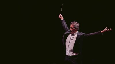 Composer Steven Mercurio, a white man with grey hair, conducts against a black background, his arms spread wide in an expressive gesture and a baton in one hand.