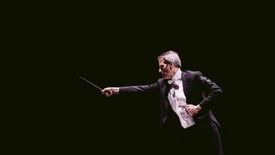 Conductor Steven Mercurio, a white man with grey hair, conducts in front of a dark background, his baton thrust forward in emphasis.