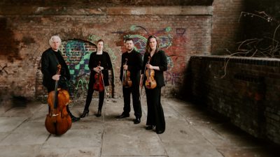 Members of the Dante Quartet hold their instruments in an industrial area of a city, in front of a brick wall covered with graffiti.