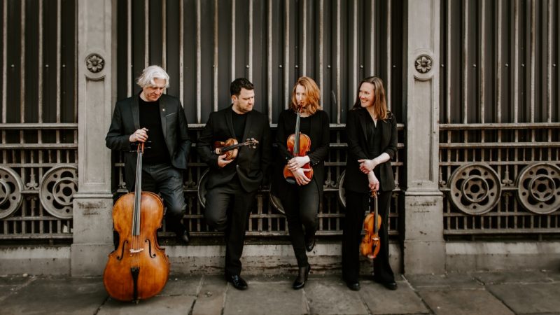 Members of the Dante Quartet hold their instruments in front of a wrought iron and stone wall in a city.