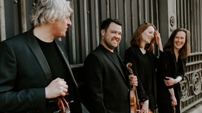 Members of the Dante Quartet hold their instruments in front of a wrought iron and stone wall in a city, and smile at each other, one man looking at the camera.