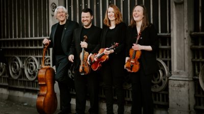 Members of the Dante Quartet hold their instruments in front of a wrought iron and stone wall in a city and laugh.