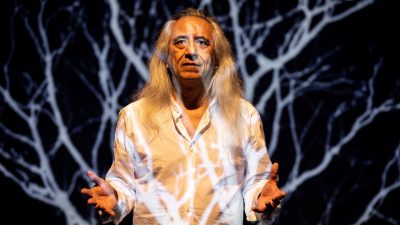 Playwright george emilio sanchez, a middle aged Indigenous man with long grey hair, wears a white button down shirt and lifts both hands in a near shrug, the projection of a desert tumbleweed behind and across him.