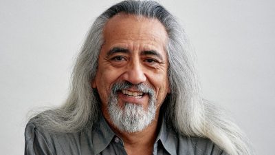 Playwright george emilio sanchez, a middle aged Indigenous man with long grey hair, wears a medium grey button down shirt and smiles towards the camera.