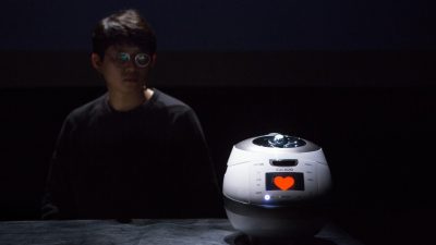 South Korean theatremaker Jaha Koo, an Asian man with short dark brown hair and round glasses, wears a black shirt and sits at a table behind a Cuckoo brand rice cooker, the screen of which displays a red heart.