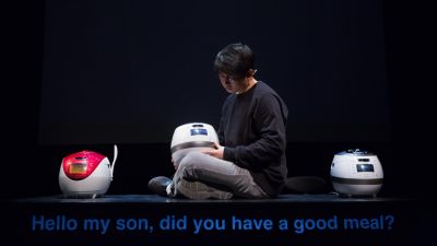 South Korean theatremaker Jaha Koo, an Asian man with short dark brown hair and round glasses, wears a black shirt and sits on top of a table between two Cuckoo brand rice cookers, another in his lap. Projected subtitles read "Hello my son, did you have a good meal?"