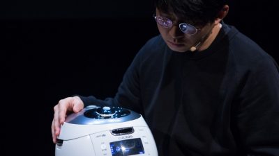 South Korean theatremaker Jaha Koo, an Asian man with short dark brown hair and round glasses, wears a black shirt and sits with a Cuckoo brand rice cooker in his lap.